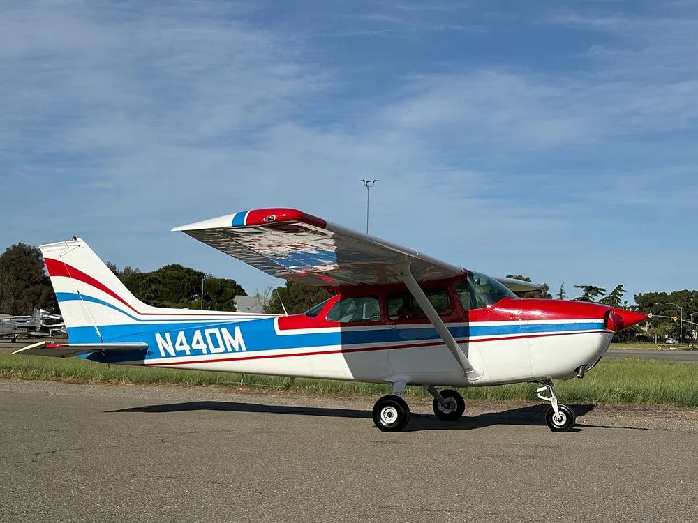 A white Cessna 172 with red and blue stripes, registration N44DM, parked on an asphalt ramp with trees and a road in the background.