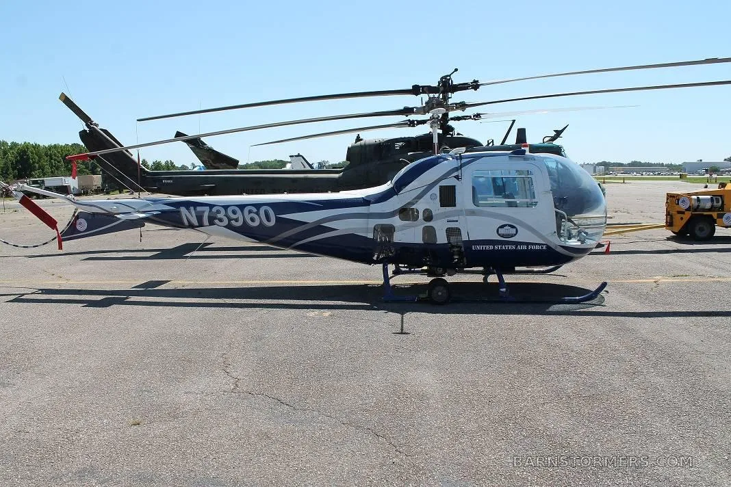 A side view of a white and blue helicopter with 'UNITED STATES AIR FORCE' text and registration N73960, parked on an airfield next to a larger dark helicopter.