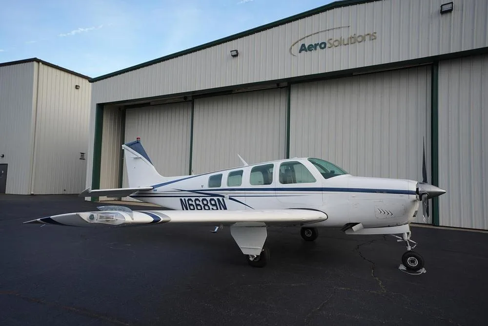 A white Beechcraft Bonanza with blue and gray stripes, registration N6689N, parked on an asphalt ramp in front of a hangar.