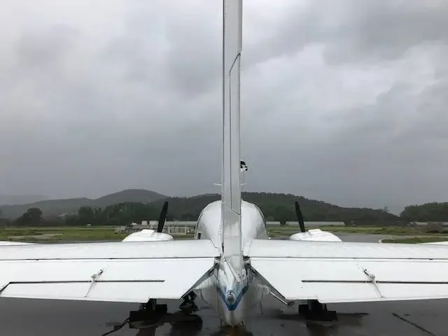 Rear view of a white twin-engine propeller aircraft on a wet tarmac with green hills in the background.