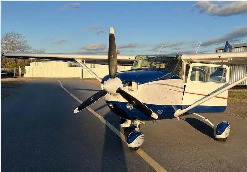 A front view of a white and blue Cessna 182, N756HC, with its propeller visible and passenger door open on an airfield.
