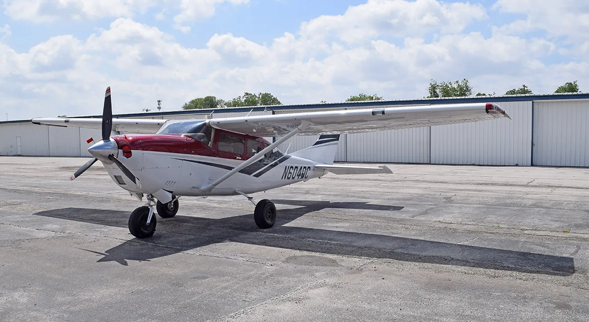 A white and red Cessna 182 aircraft, registration N604DC, parked on an asphalt tarmac with hangars in the background.