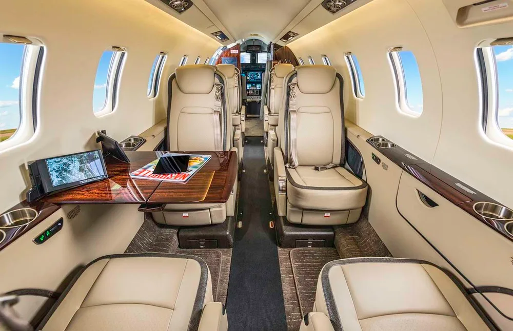 The luxurious interior cabin of a private jet features beige leather seats, a polished wooden table, and large windows.