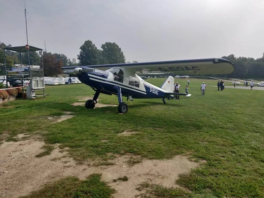 A blue and white high-wing aircraft, registration D-EOAC, parked on a grassy airfield with people and other aircraft in the background.