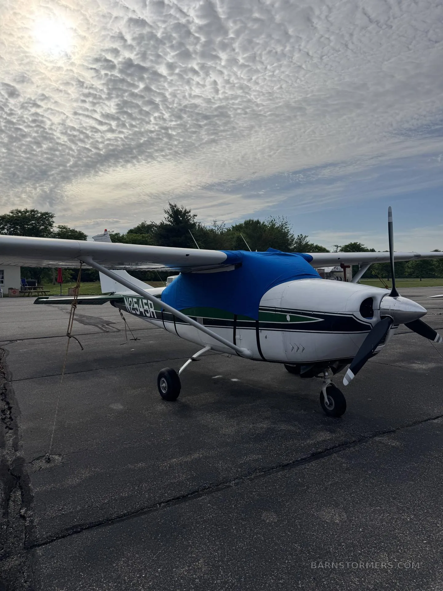 A white and green single-engine propeller aircraft with a blue engine cover is parked on an asphalt ramp under a cloudy sky.