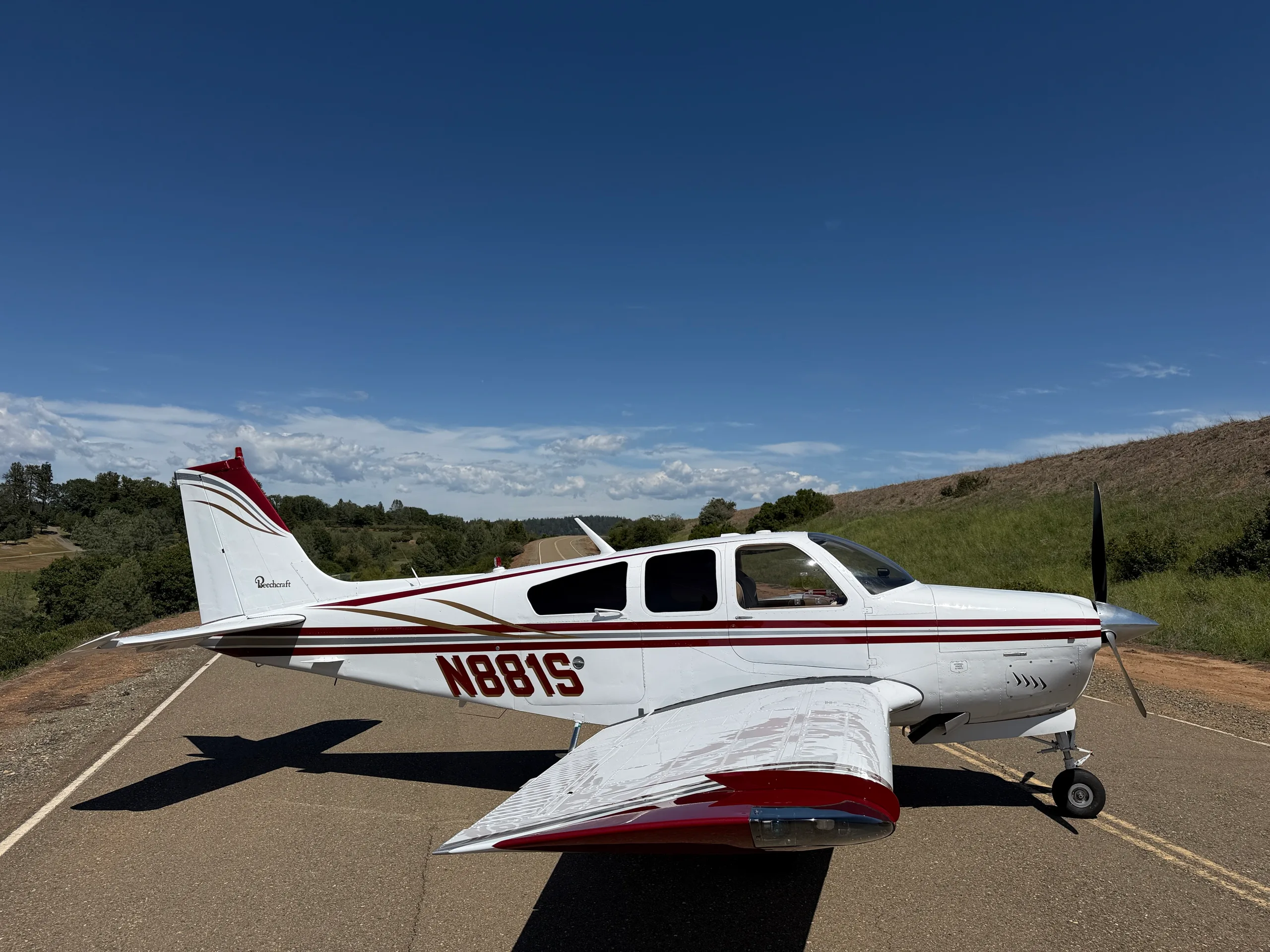 A white Beechcraft Bonanza N881S with red and gold stripes is parked on a paved surface under a clear blue sky.