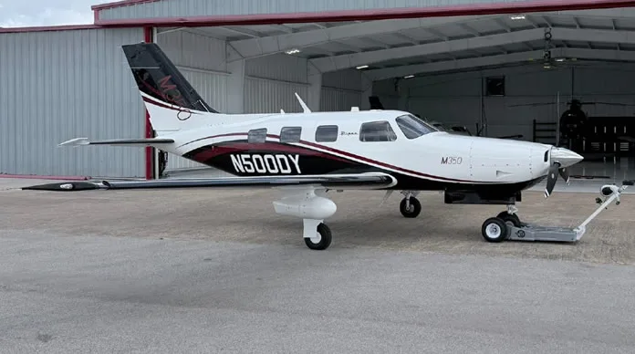 A white Piper M350 aircraft with black and maroon stripes is parked on the tarmac outside an open hangar.