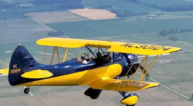 A blue and yellow biplane with registration NC 30124 flies over a patchwork of green and brown fields.