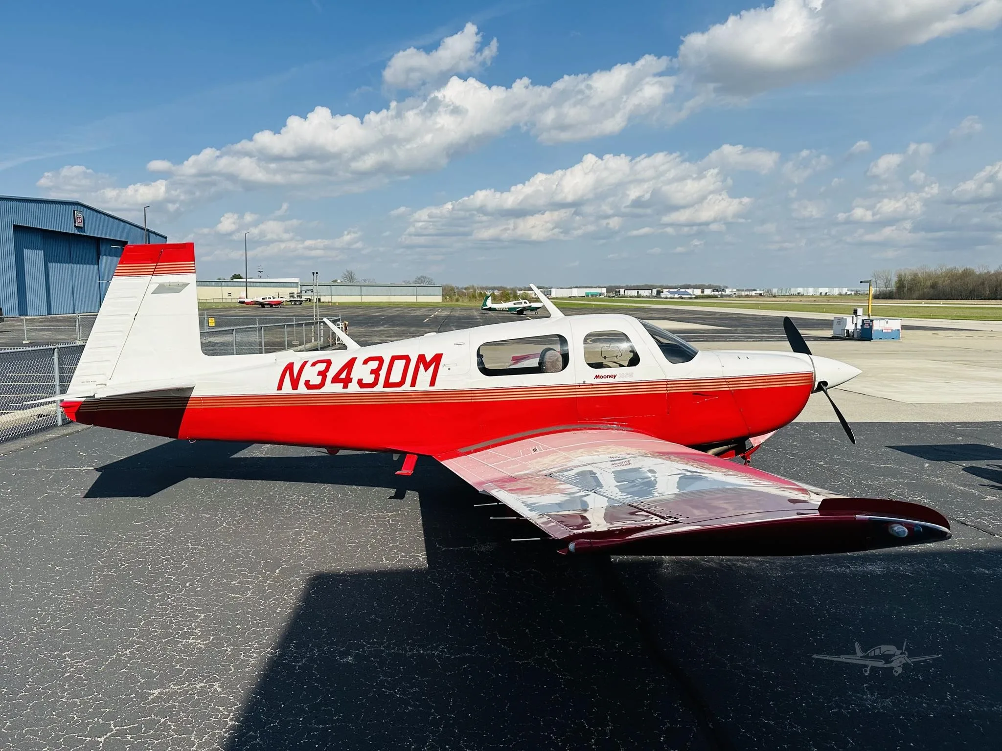 A red and white Mooney M20 aircraft with registration N343DM parked on an asphalt tarmac under a partly cloudy sky.