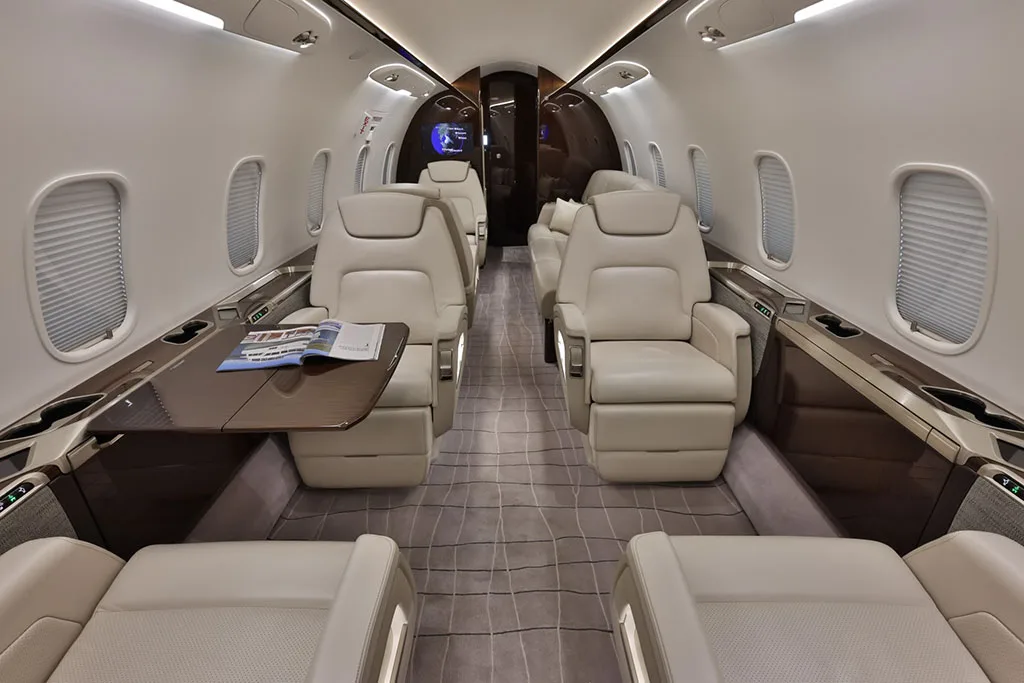 The luxurious interior cabin of a private jet, featuring beige leather seats, a dark wood table, and a patterned carpet.