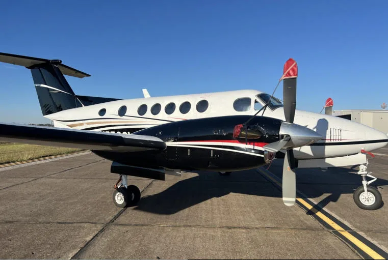 A Beechcraft King Air 350 with a black and white paint scheme and gold accents, parked on an airfield under a clear blue sky.