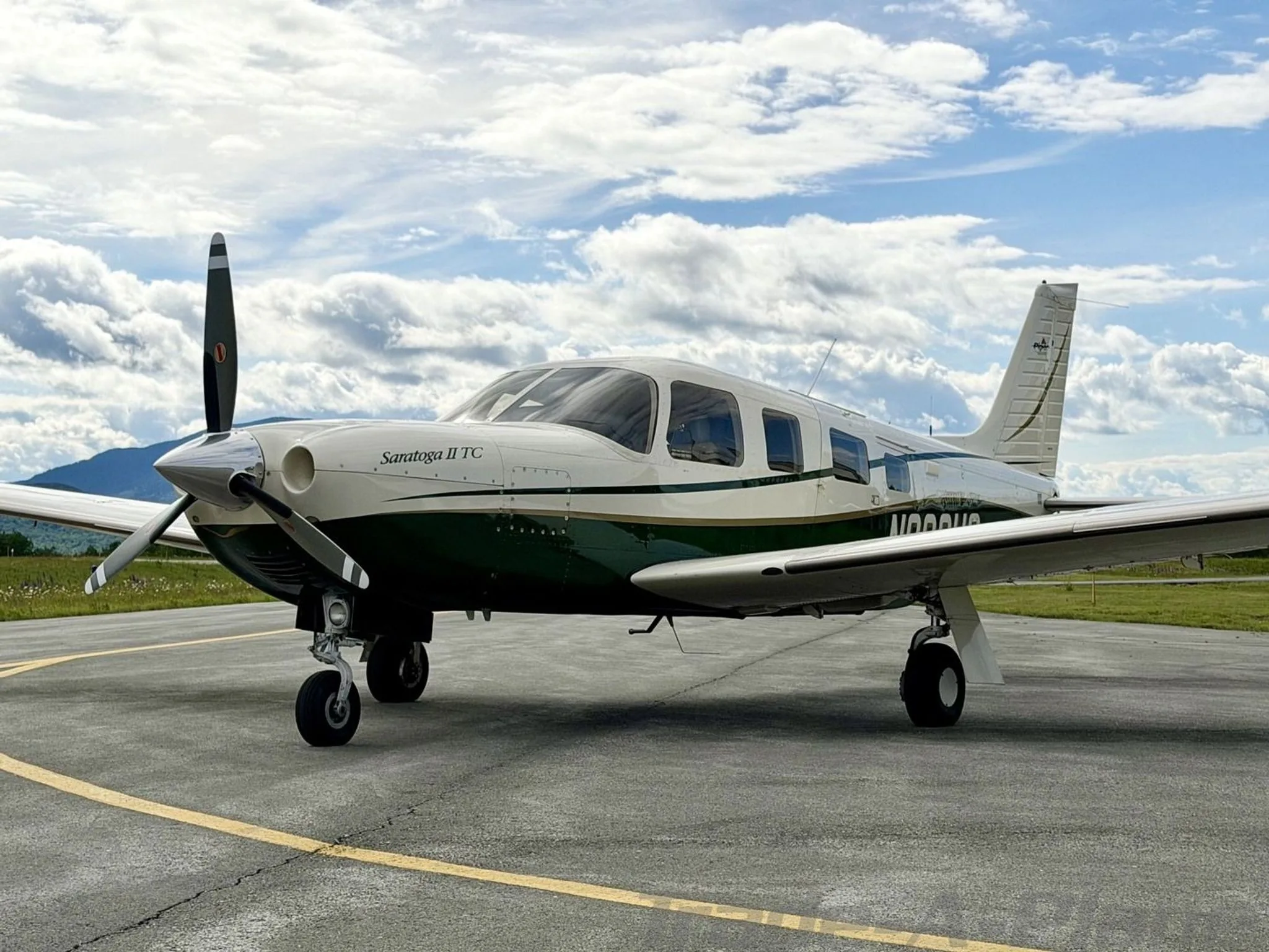 A white and green Piper Saratoga II TC aircraft, registration N800HS, parked on an asphalt ramp with mountains in the background under a partly cloudy sky.