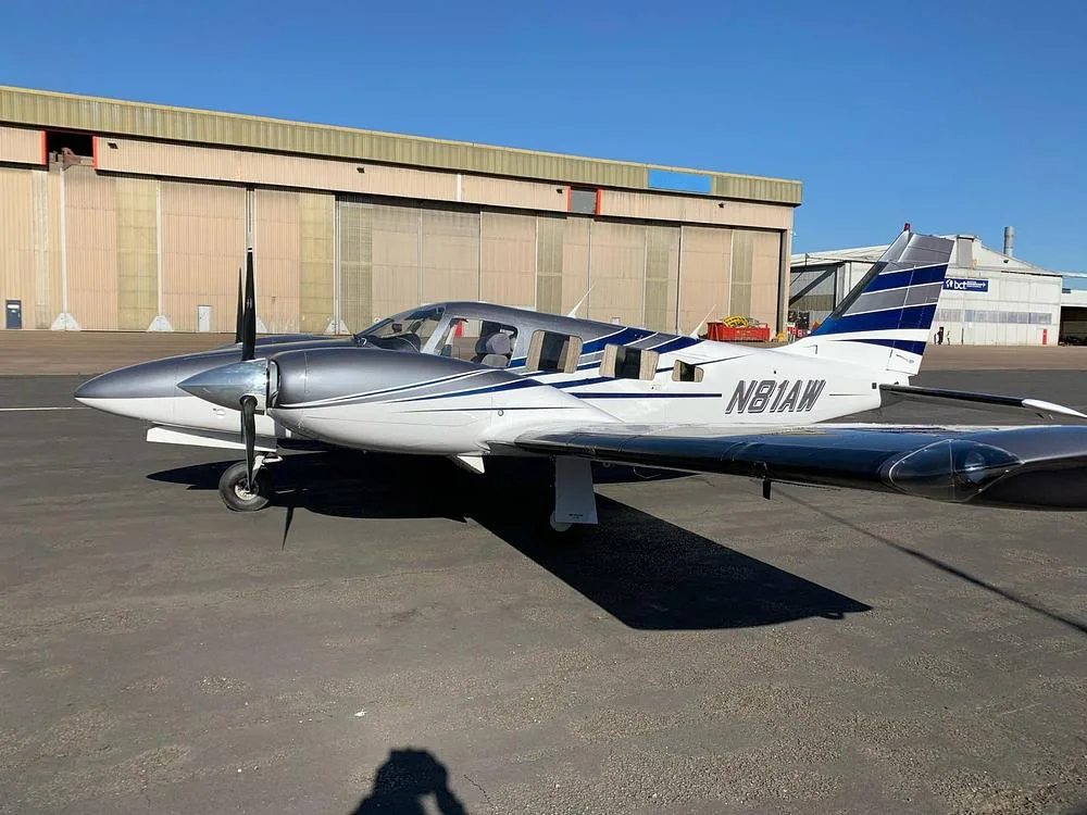 A white and grey twin-engine aircraft (N81AW) with blue stripes parked on an airfield next to a large hangar.