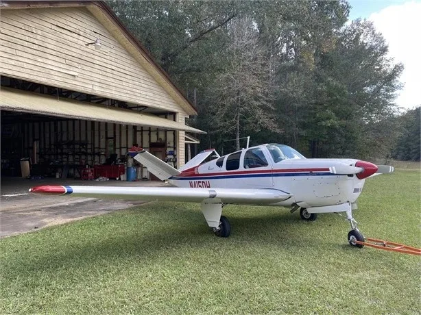 A white and red Beechcraft Bonanza N415DH parked on grass with its nose facing a wooden hangar.