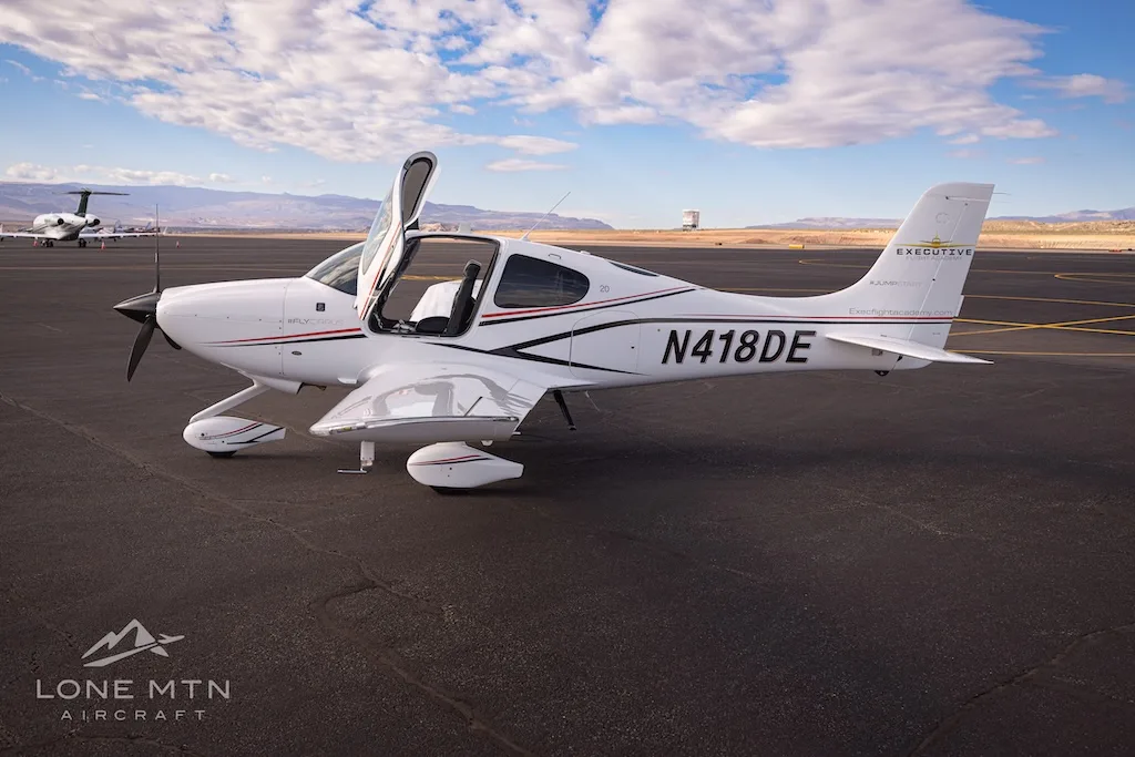 A white Cirrus SR22 aircraft with black and red stripes, registration N418DE, parked on an airfield with doors open.