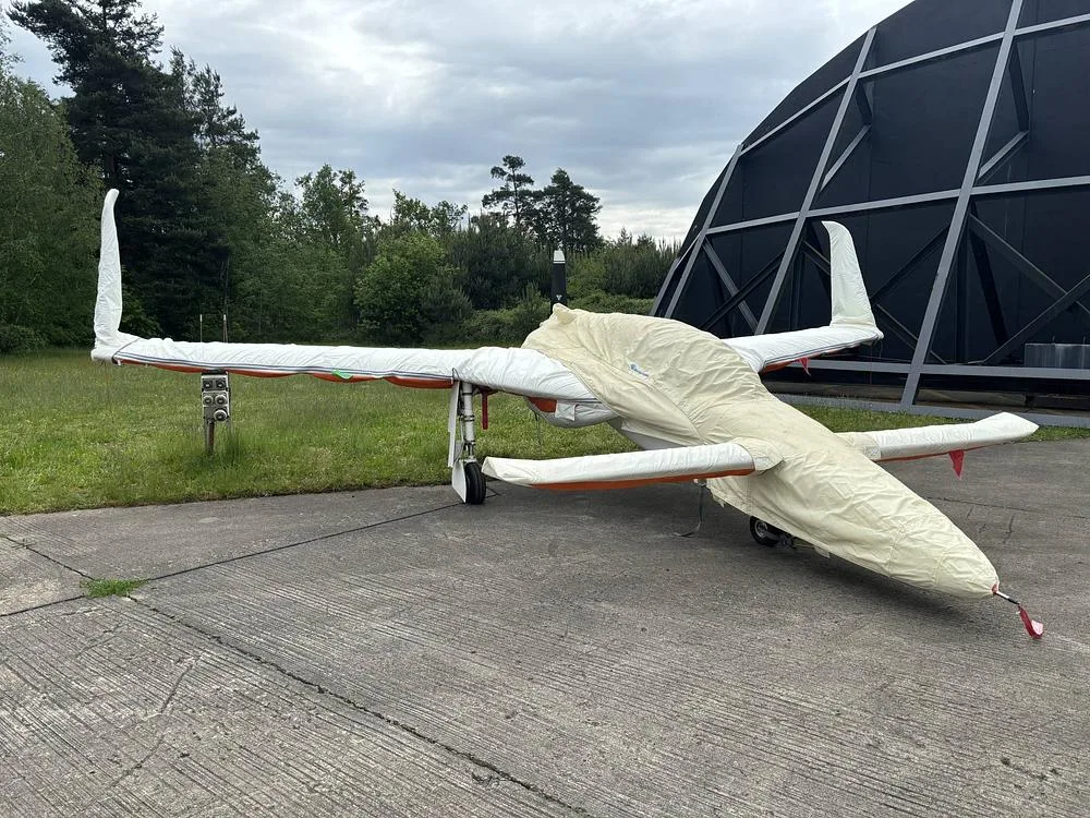 A white, single-engine aircraft, partially covered, is parked on a concrete pad next to a dark dome-shaped building and trees.