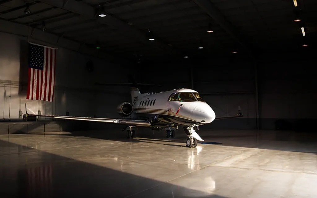 A white and black private jet with gold stripes parked in a dimly lit hangar with an American flag hanging in the background.