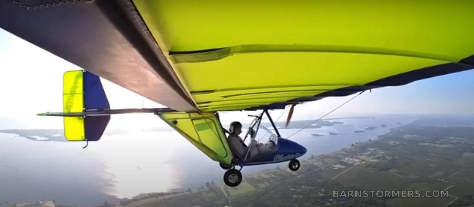A pilot's view from a blue ultralight cockpit, showing instruments and a chicken in the pilot's lap, flying over green terrain.
