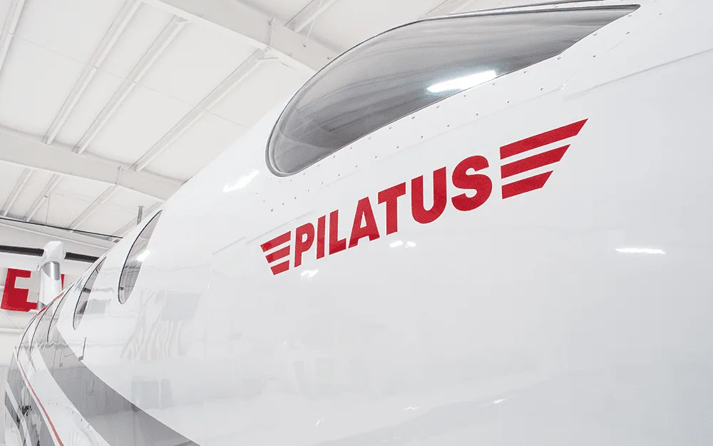 A close-up view of the white fuselage of a Pilatus PC-12 showing the red "PILATUS" logo and cockpit window.