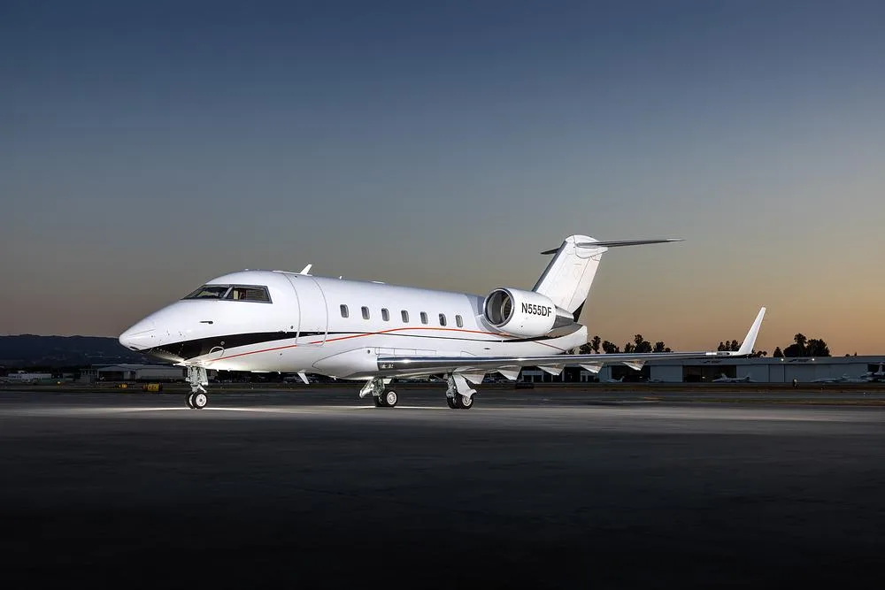A white Bombardier Challenger 605 business jet, registration N555DF, sits on an airport tarmac at sunset.