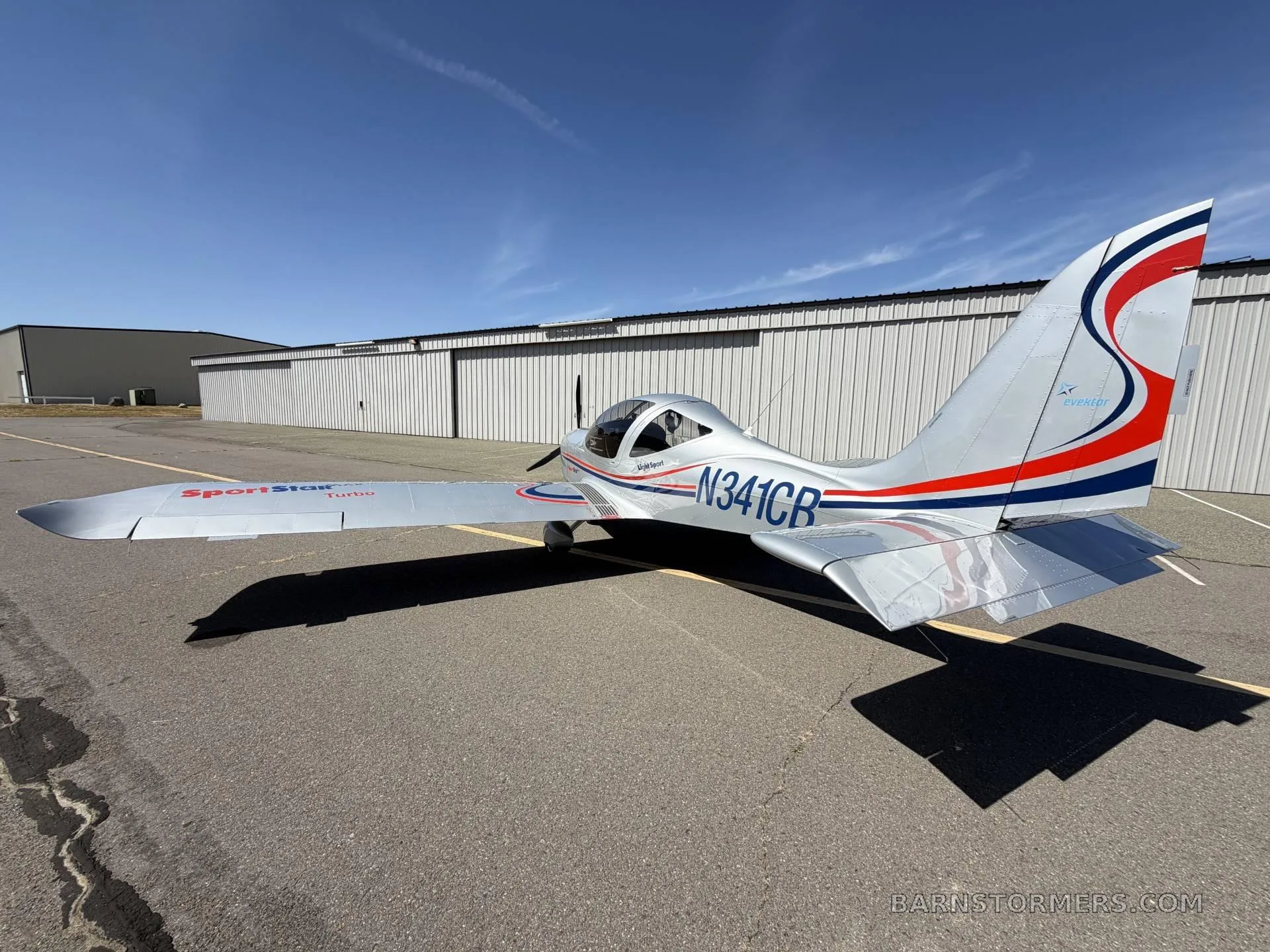 A rear-left view of the silver SportStar MAX Turbo aircraft with tail number N341CB and red and blue stripes, parked on an asphalt tarmac.