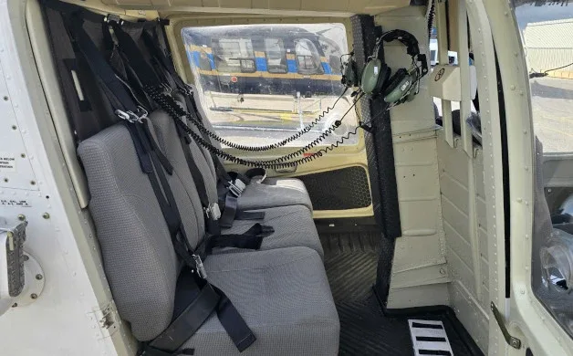 The interior cabin of a helicopter showing three grey passenger seats with harnesses and a headset hanging on the wall.