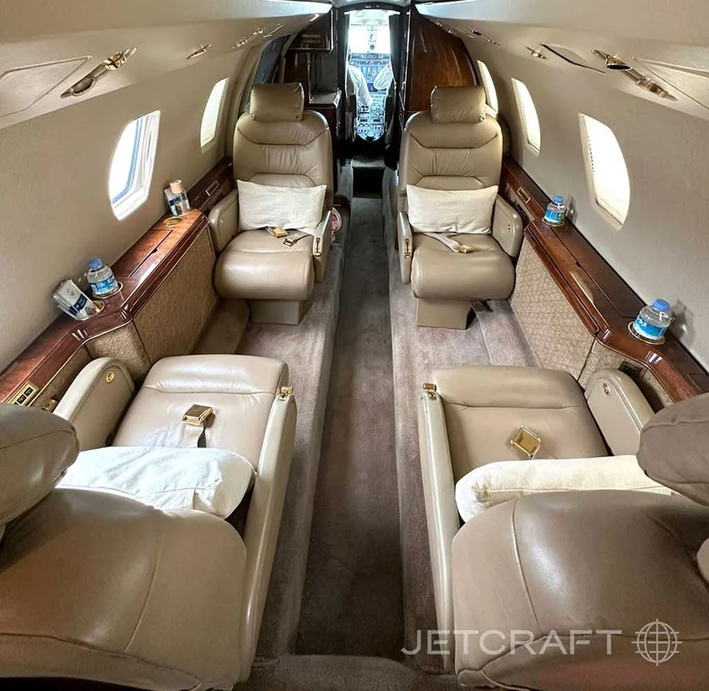 The luxurious interior cabin of a private jet, featuring tan leather seats and wood trim, looking towards the cockpit.