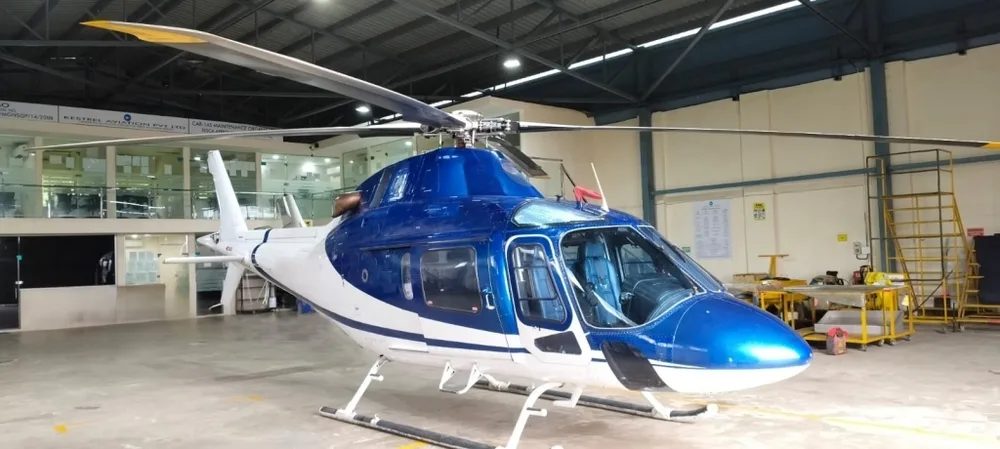 A front-quarter view of a blue and white helicopter with its main rotor blades extended, inside a hangar.