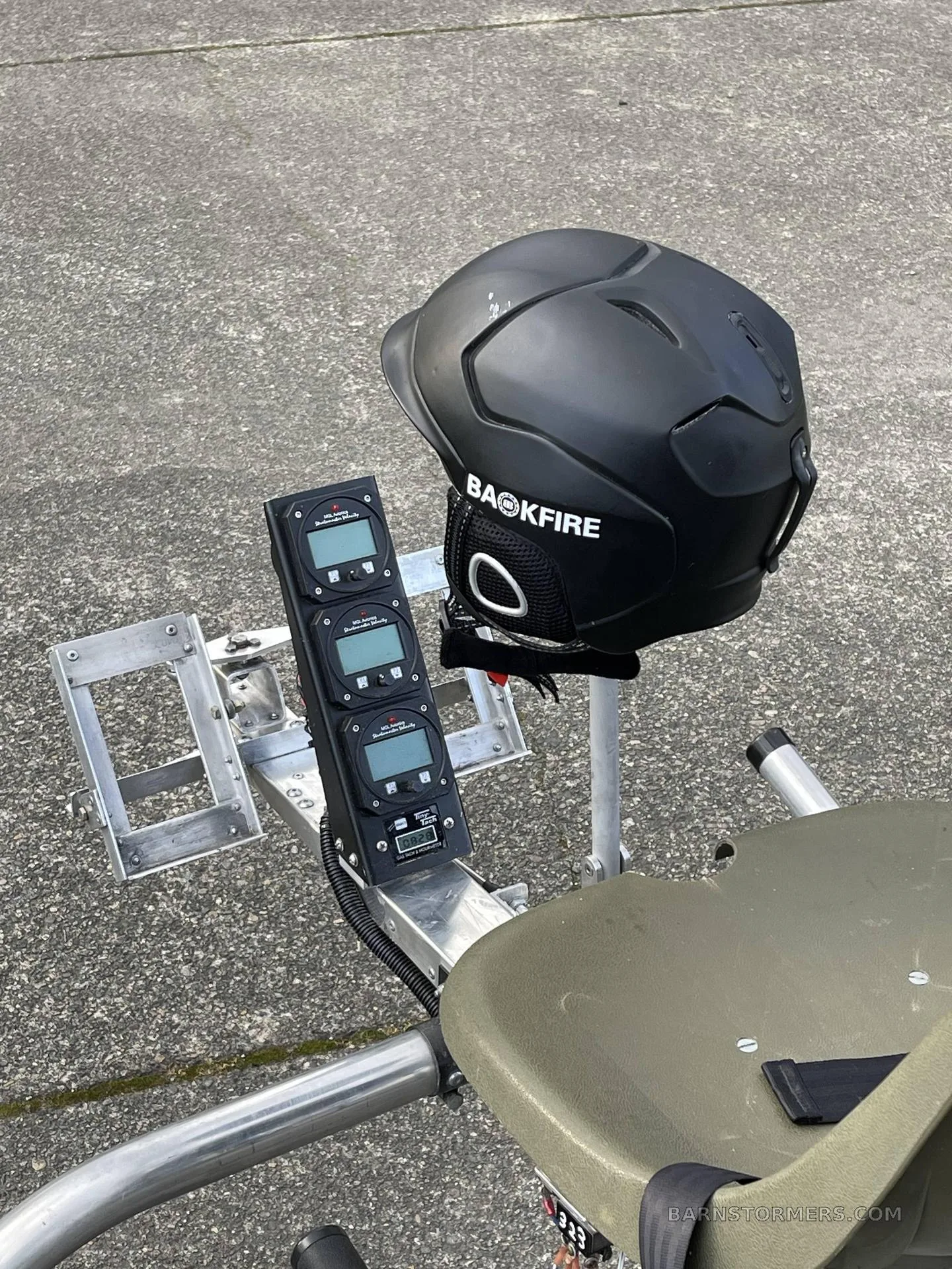 A close-up view of the minimalist instrument panel with three digital gauges and a black "BACKFIRE" helmet resting on the seat of an ultralight helicopter.