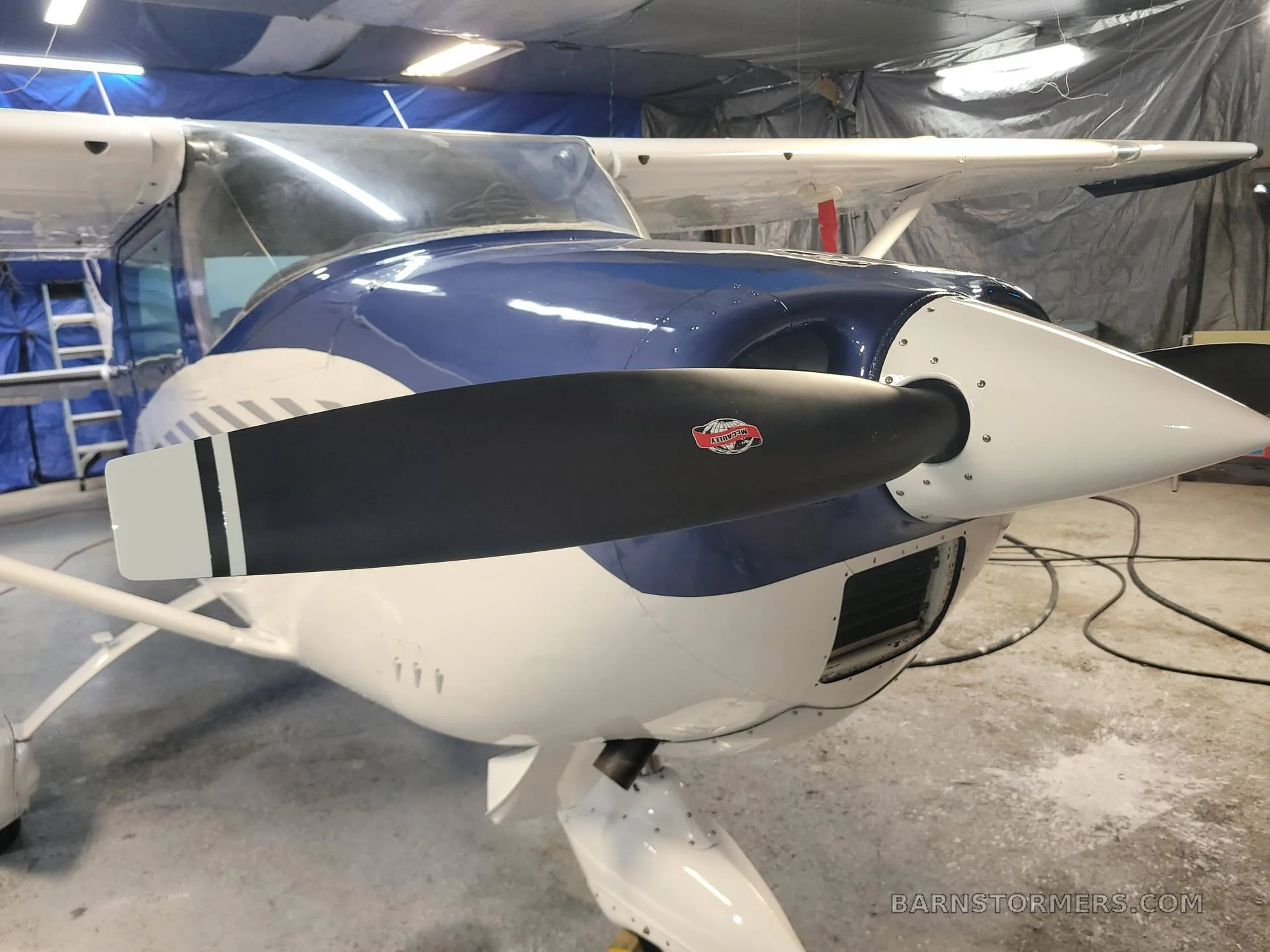 Front view of a blue and white aircraft, showing the propeller and nose cone in a hangar.
