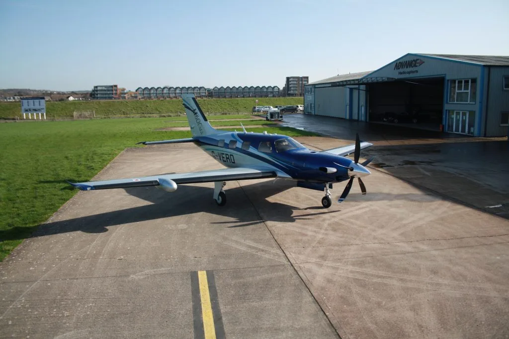 A blue and silver Piper M600SLS aircraft (G-VERO) parked on a tarmac next to an "Advance Helicopters" hangar.