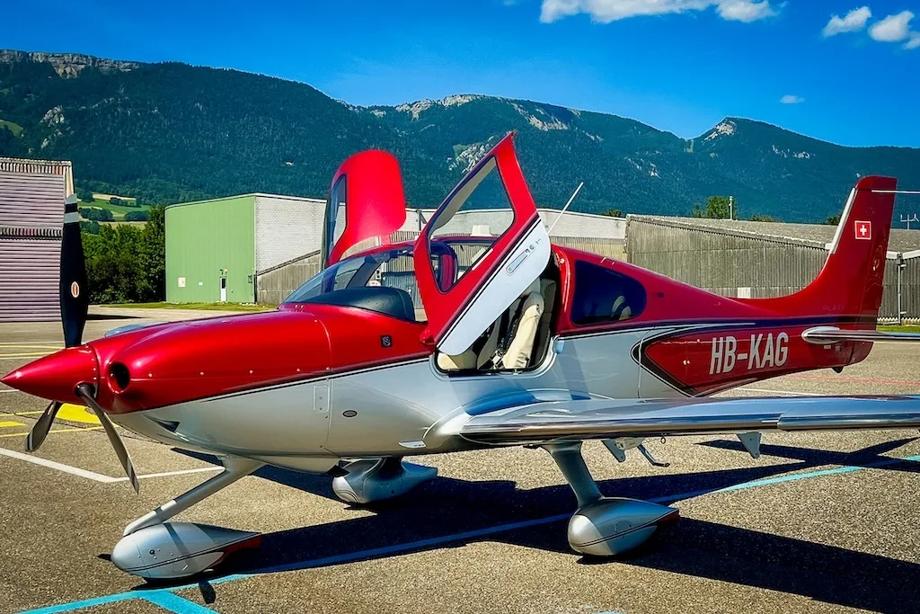 Red and silver Cirrus SR22 aircraft with doors open, parked on an airfield with mountains in the background.