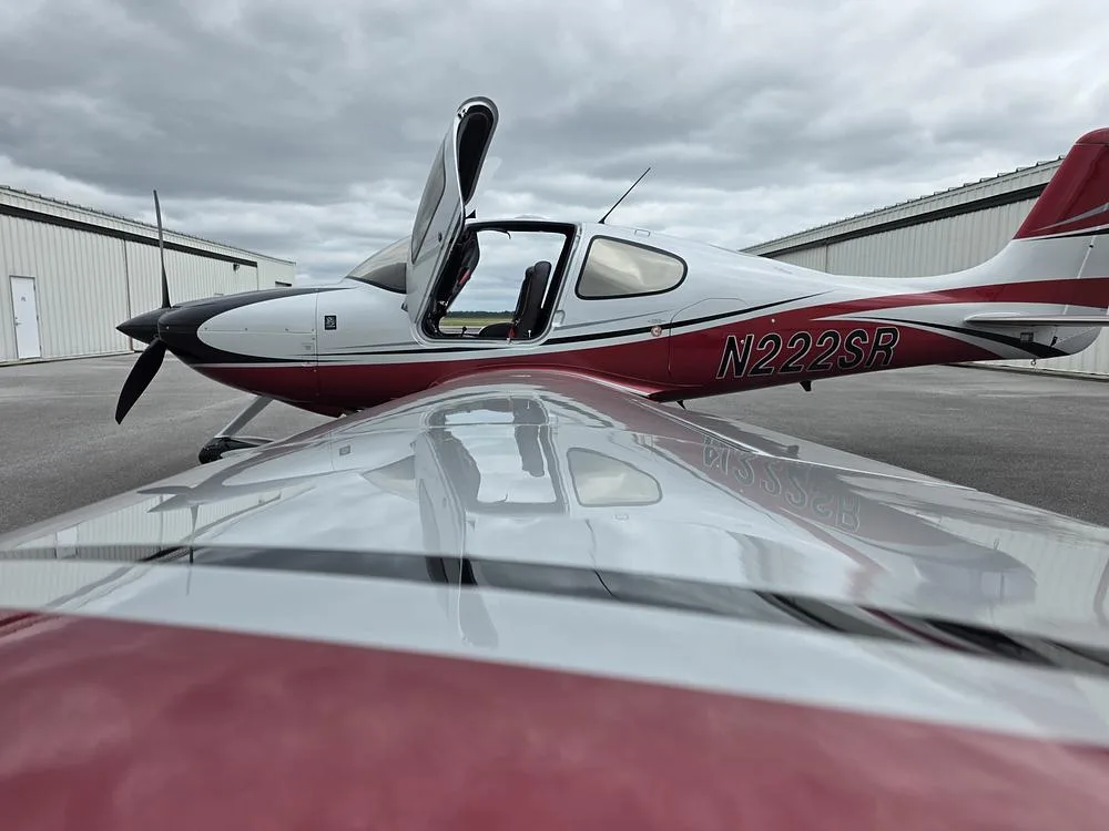 A white, red, and black single-engine aircraft with open doors, viewed from a low angle over the wing.