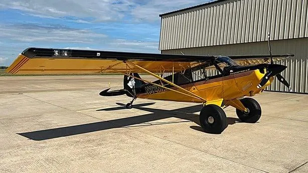 A yellow and black Aviat Husky A-1C aircraft with large tundra tires is parked on a concrete tarmac in front of a hangar, viewed from the front-left.