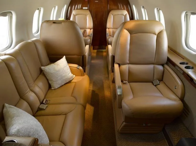 A view of the private jet cabin interior with tan leather seats, including a side couch and individual chairs, and decorative pillows.