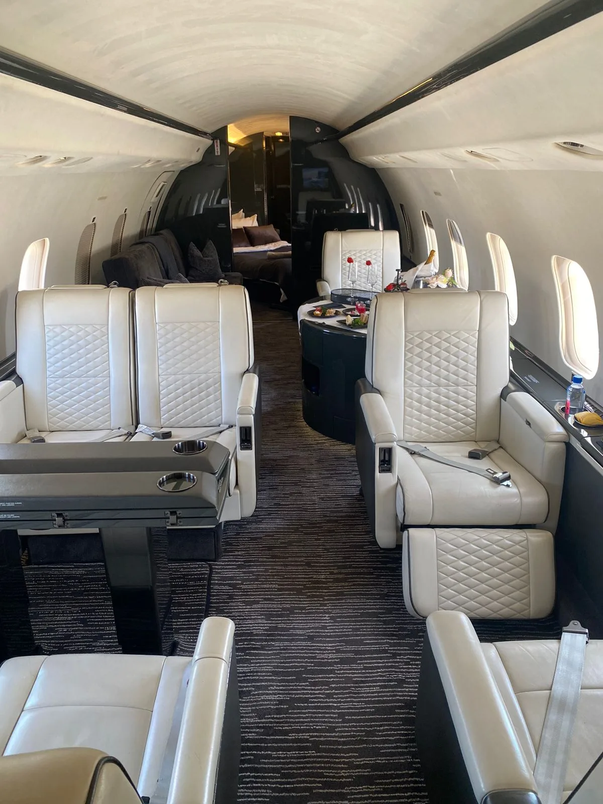The spacious interior of a private jet featuring white quilted leather seats, a dining area, and a rear lounge.