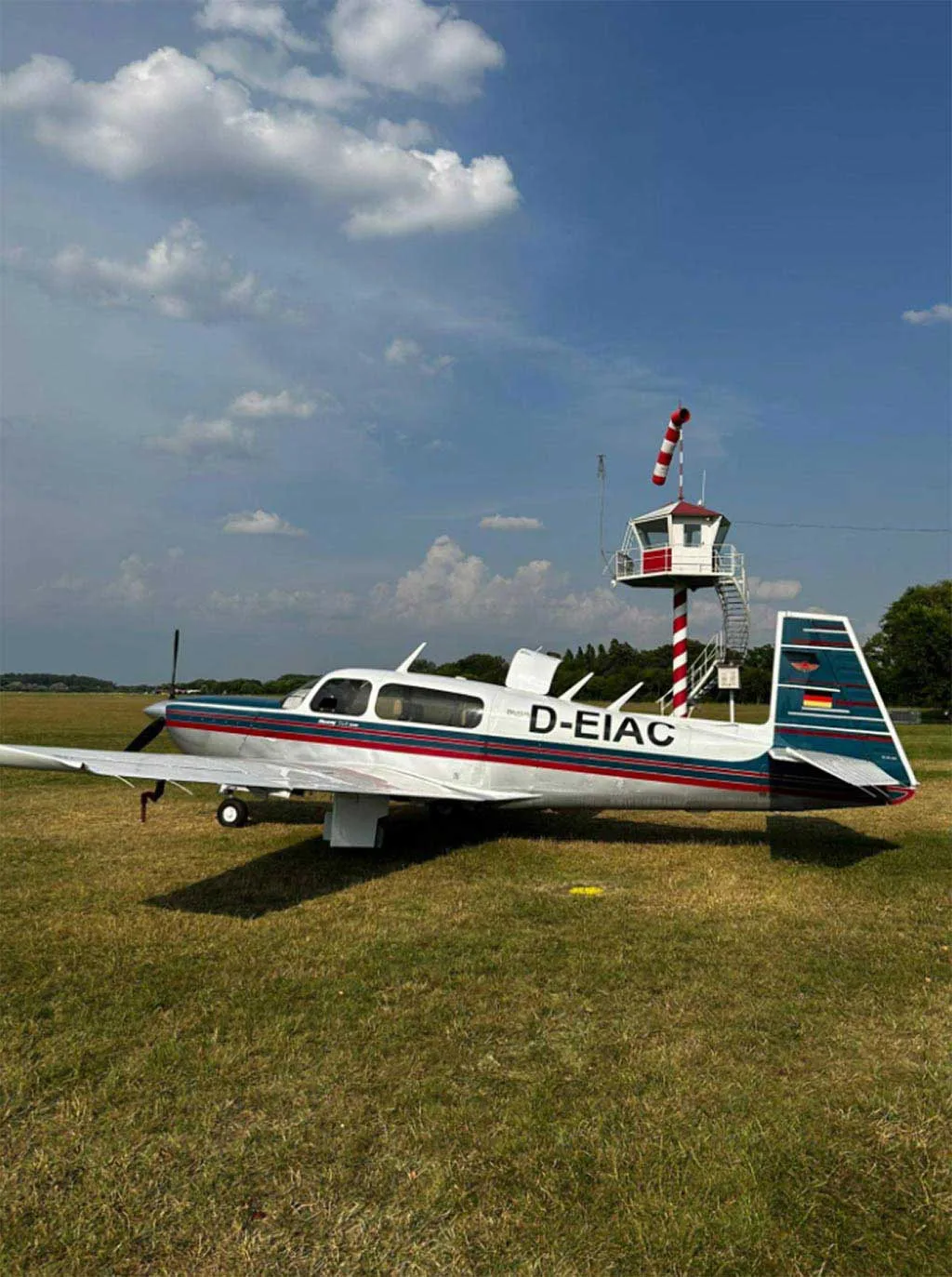 A Mooney M20J aircraft with a silver, blue, and red paint scheme parked on a grassy field next to a small red and white striped control tower.
