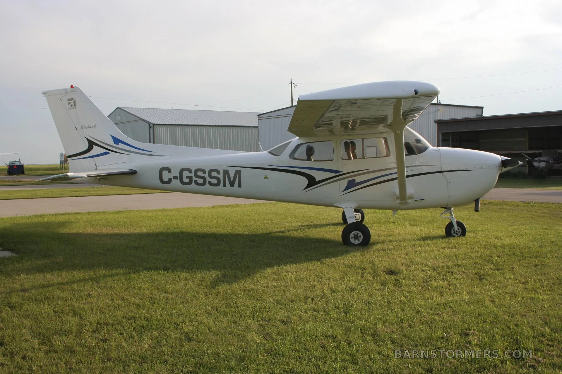 A white Cessna 172 (C-GSSM) with blue and black stripes is parked on green grass next to hangars.
