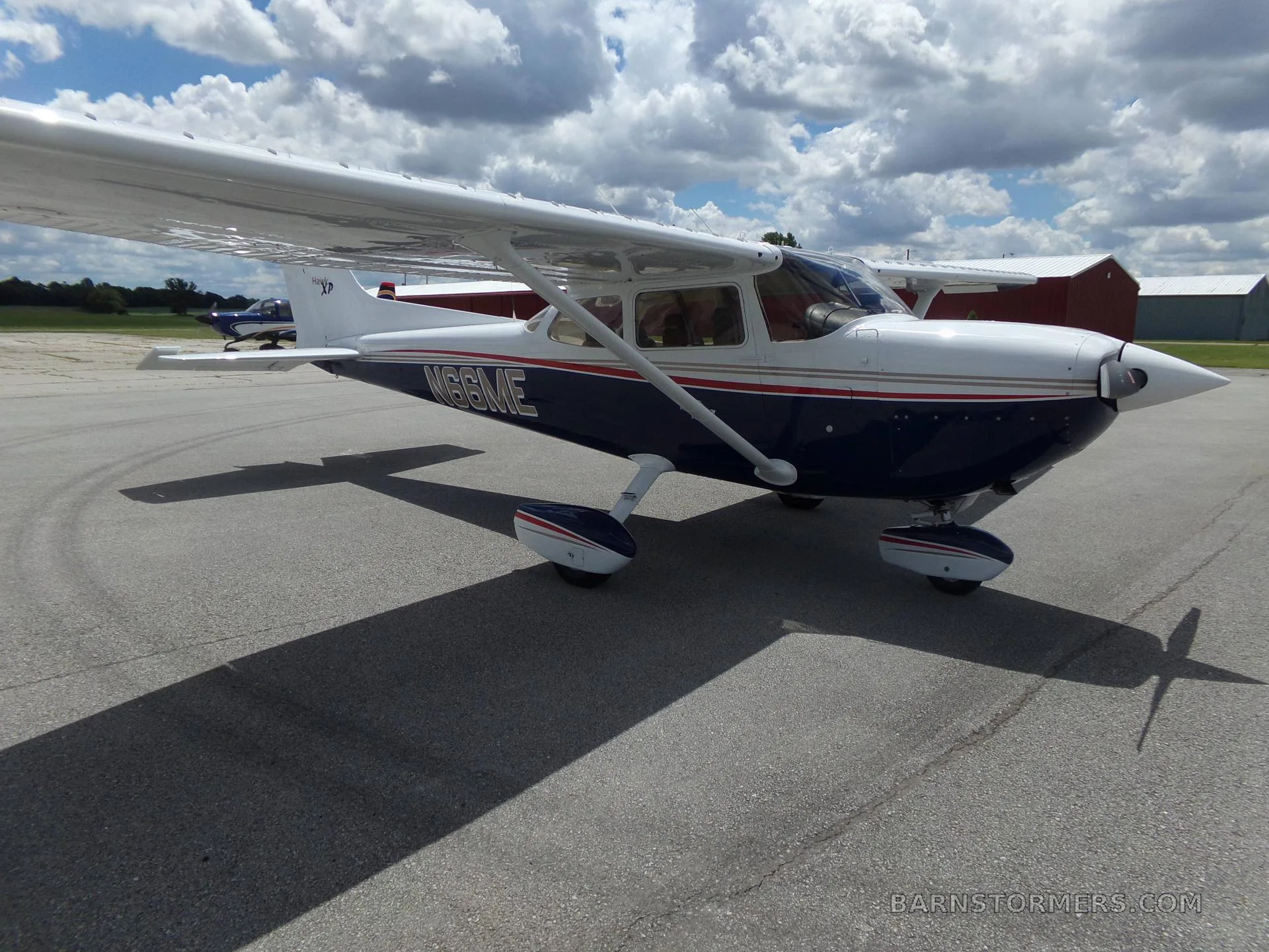 A white and blue Cessna 172 Hawk XP with red and gold stripes, seen from the front-right on an airfield with hangars in the background.