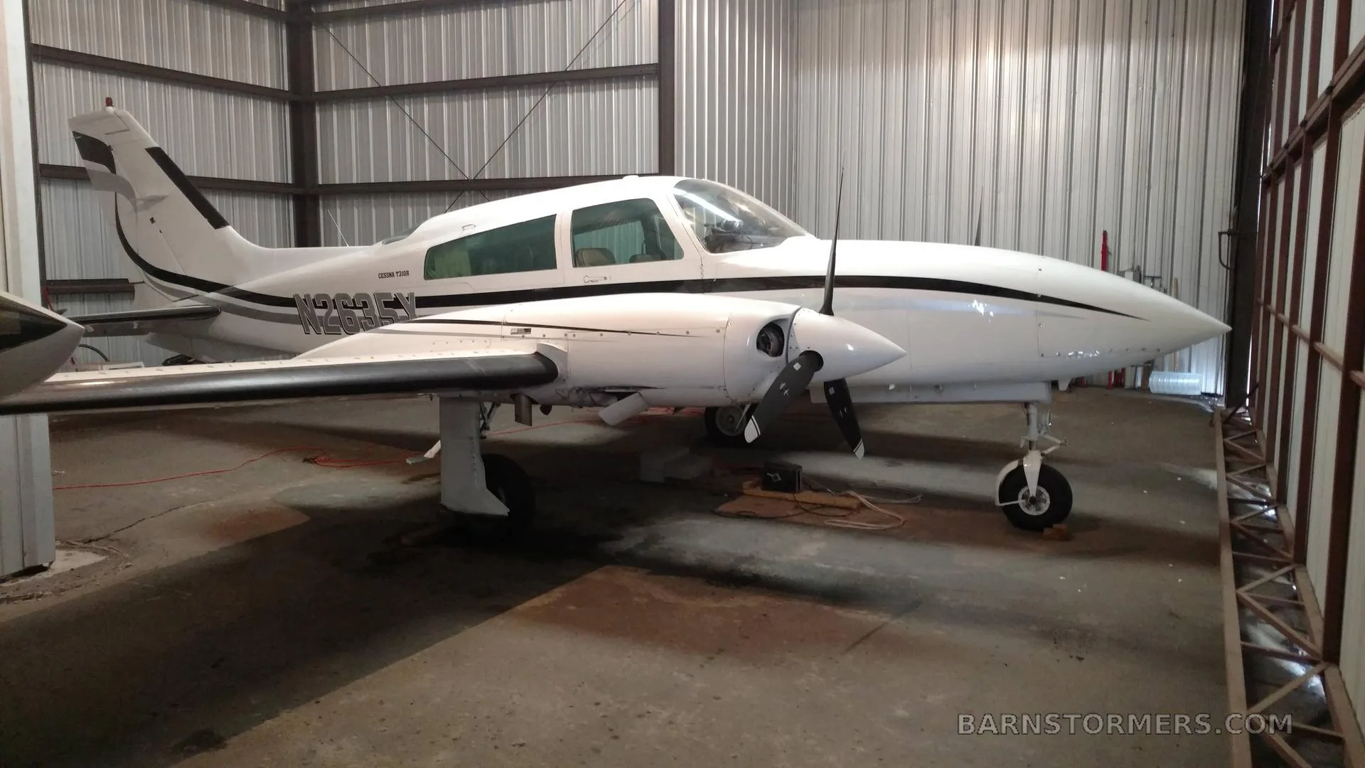 A white Cessna 310 twin-engine aircraft with black stripes is parked inside a metal hangar, viewed from the front-right.