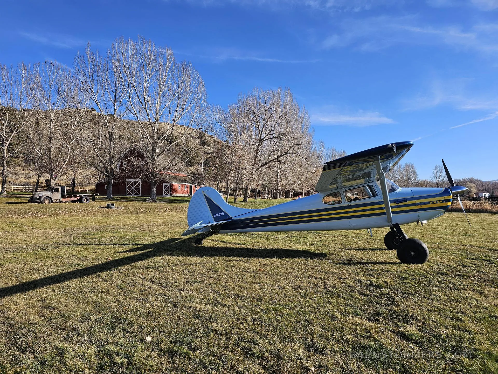 A light blue and yellow striped taildragger aircraft is parked in a grassy field with a red barn and trees in the background.