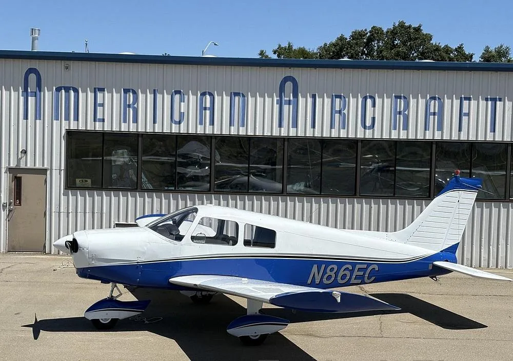 A white and blue Piper Cherokee aircraft (N86EC) parked in front of a building labeled "AMERICAN AIRCRAFT".