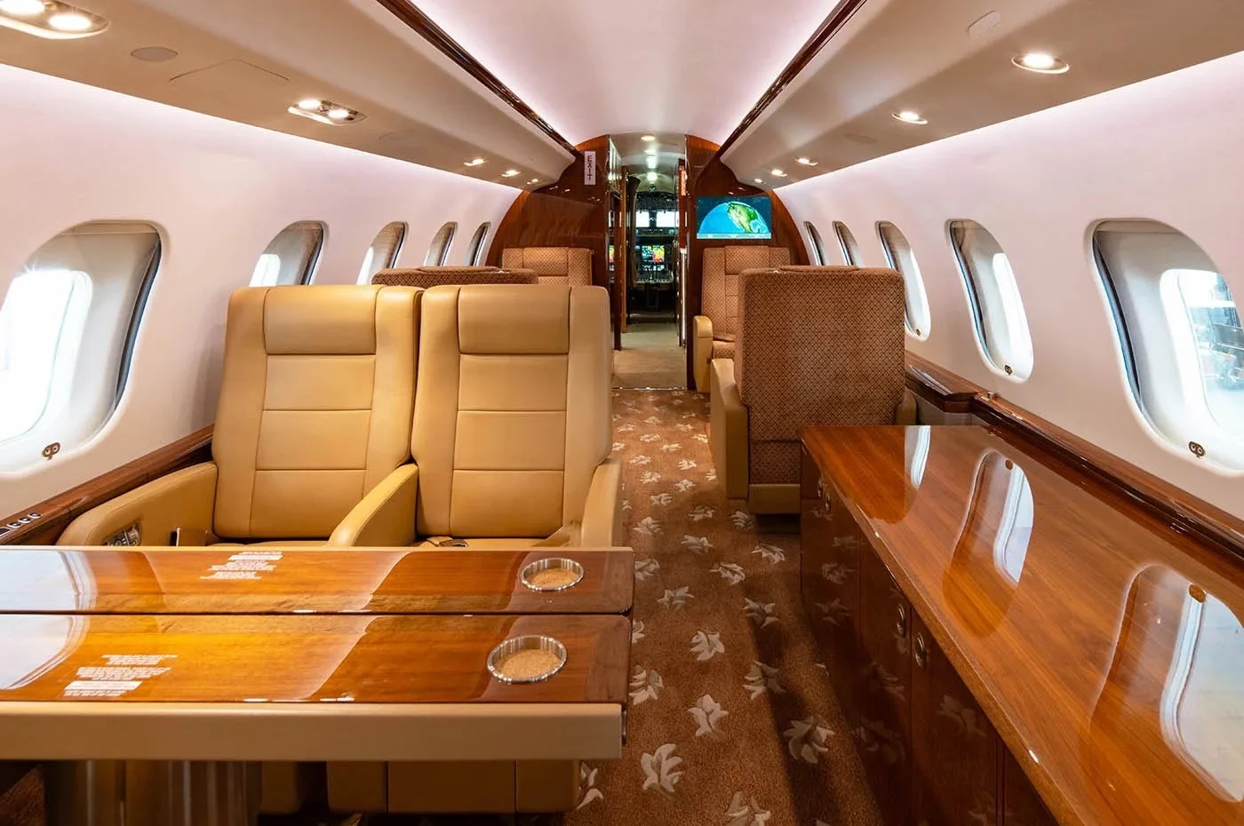 The luxurious aircraft cabin features tan leather seats, wooden tables, and a patterned carpet.