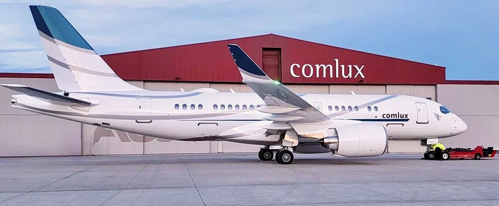 A white Comlux business jet with blue and grey tail accents parked on an airfield in front of a red hangar.