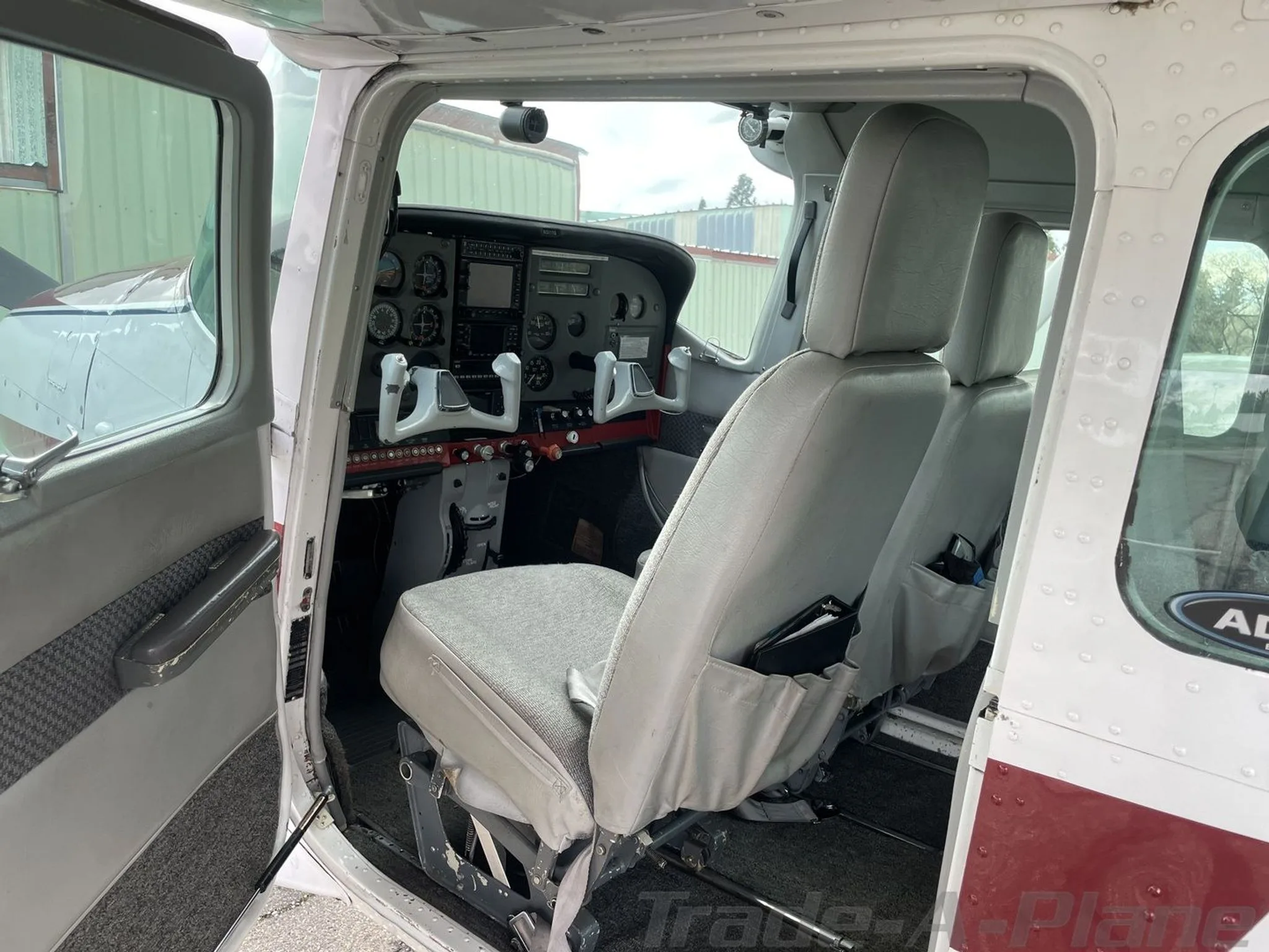 The open door view of a Cessna 182 aircraft interior, showing gray fabric seats and a partial view of the cockpit.
