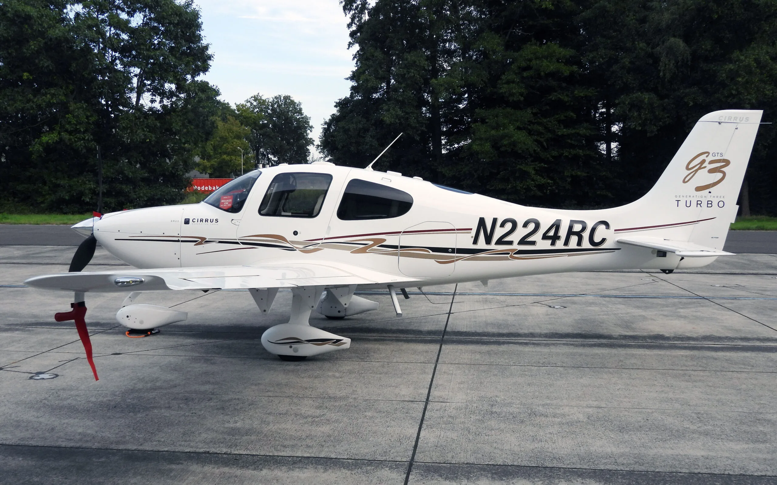A white Cirrus SR22T (N224RC) with brown and black stripes parked on an asphalt apron with trees in the background.
