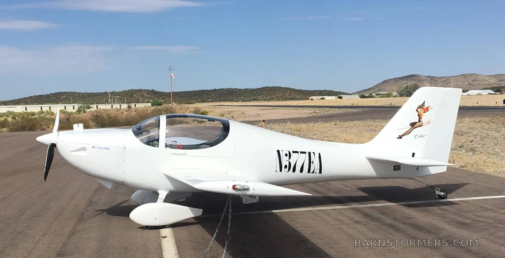 A white single-engine aircraft with "N377EA" on the side and a pin-up girl on the tail, parked on an airfield.