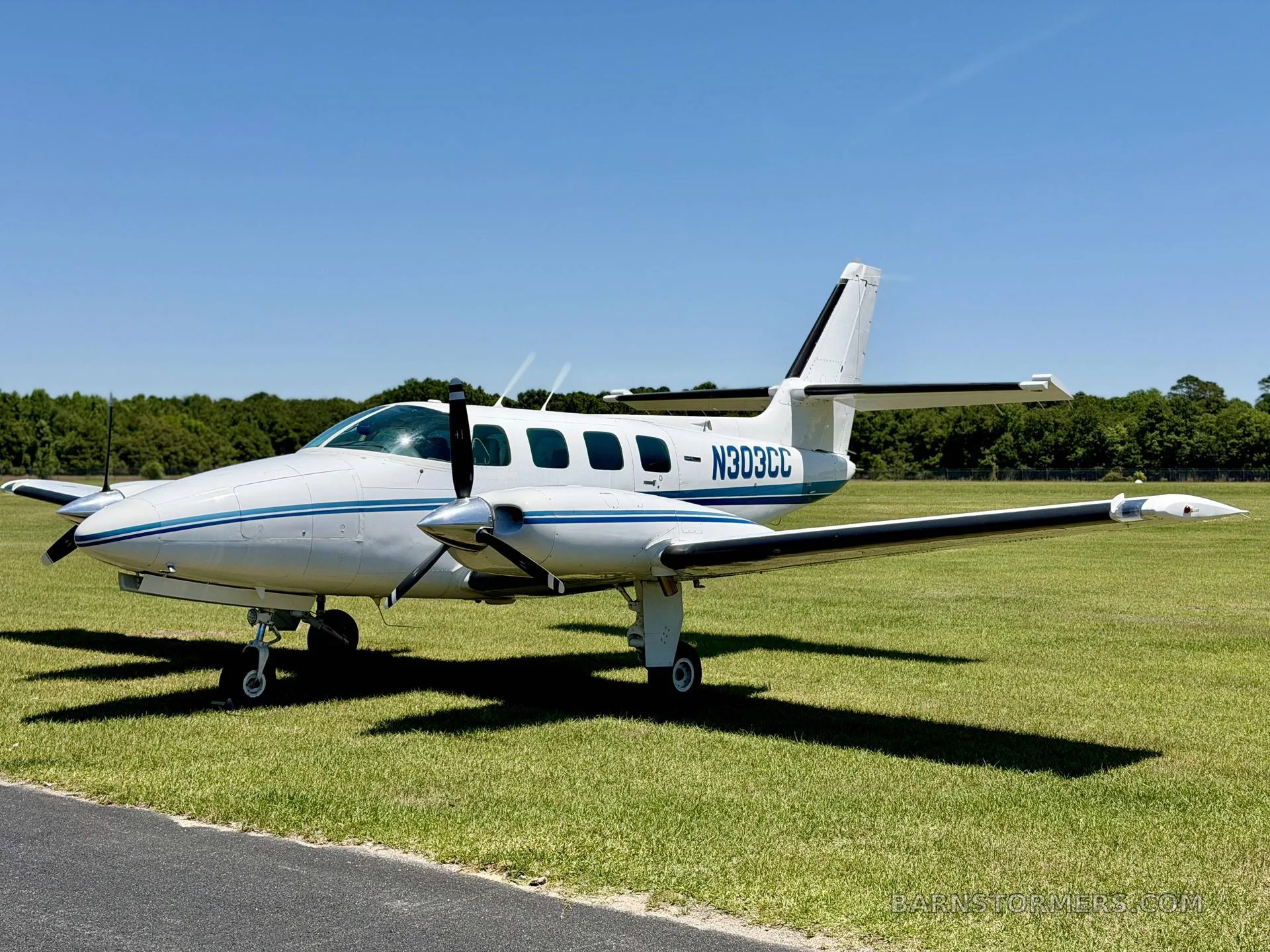 A white twin-engine propeller aircraft with blue stripes (N303CC) is parked on a grassy field under a clear blue sky.