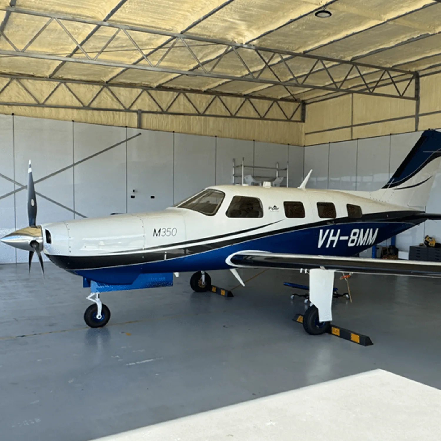 A white and blue Piper M350 aircraft with registration VH-8MM parked inside a hangar.