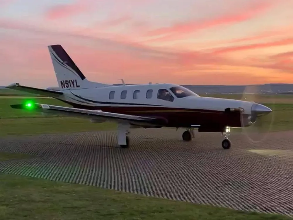 Side view of a white and maroon single-engine turboprop aircraft with its propeller spinning, on a runway at sunset.